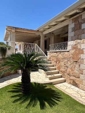 A stone building with a covered porch, a palm tree, and a staircase leading to a door.
