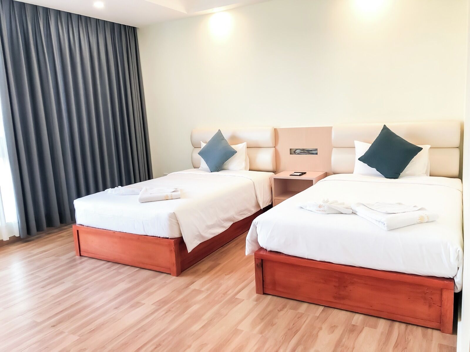 Een hotelkamer met twee bedden, houten vloeren, een nachtkastje en een groot raam met gordijnen.