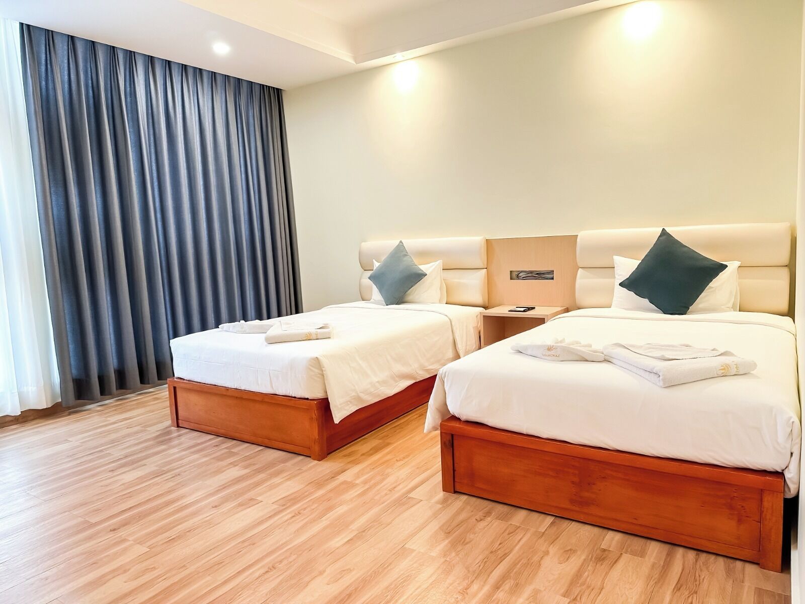Habitación de hotel con dos camas, piso de madera, una mesita de noche y un ventanal con cortinas.