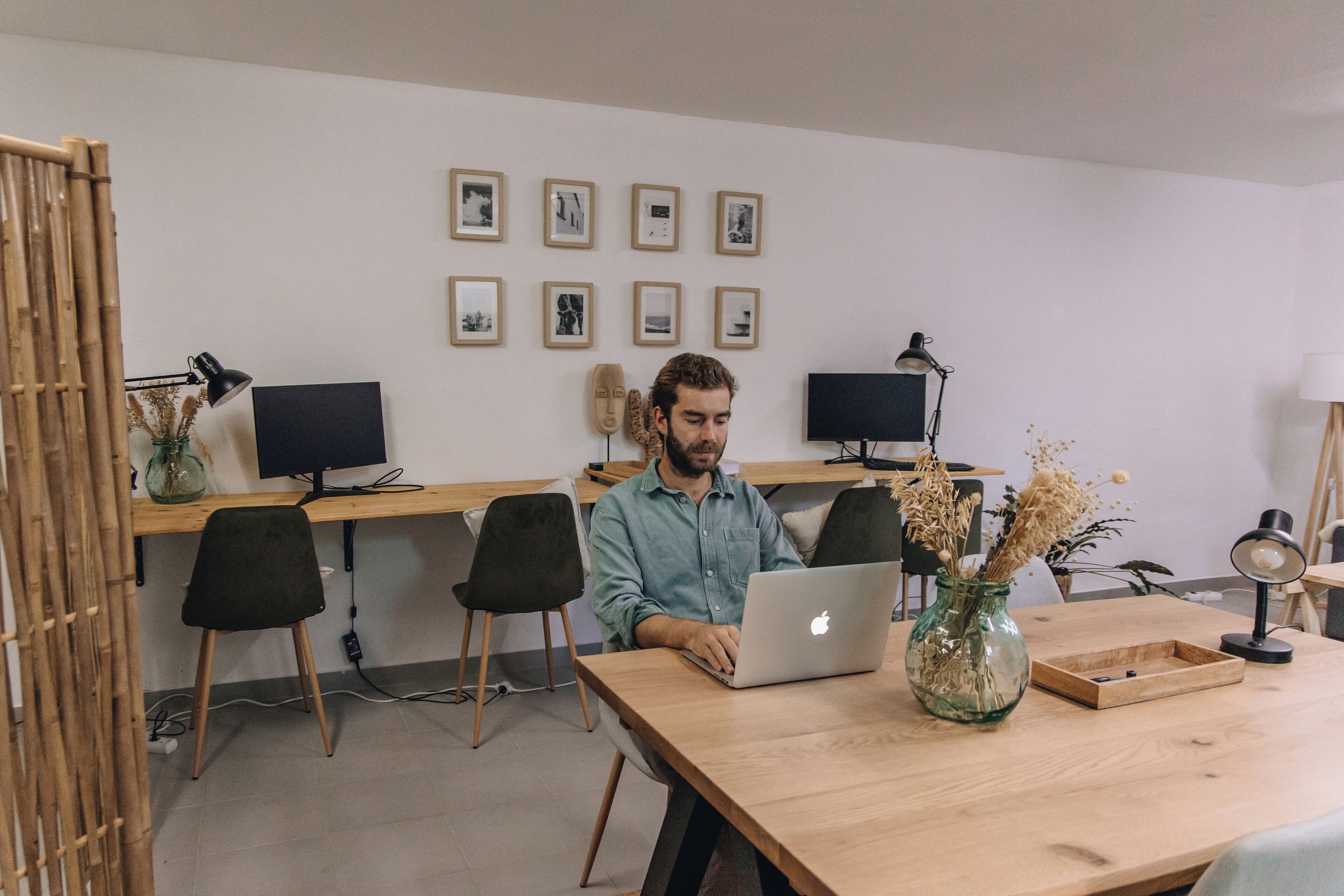 Una stanza con una scrivania in legno, due monitor per computer, un laptop, un vaso con fiori secchi e quadri appesi al muro.