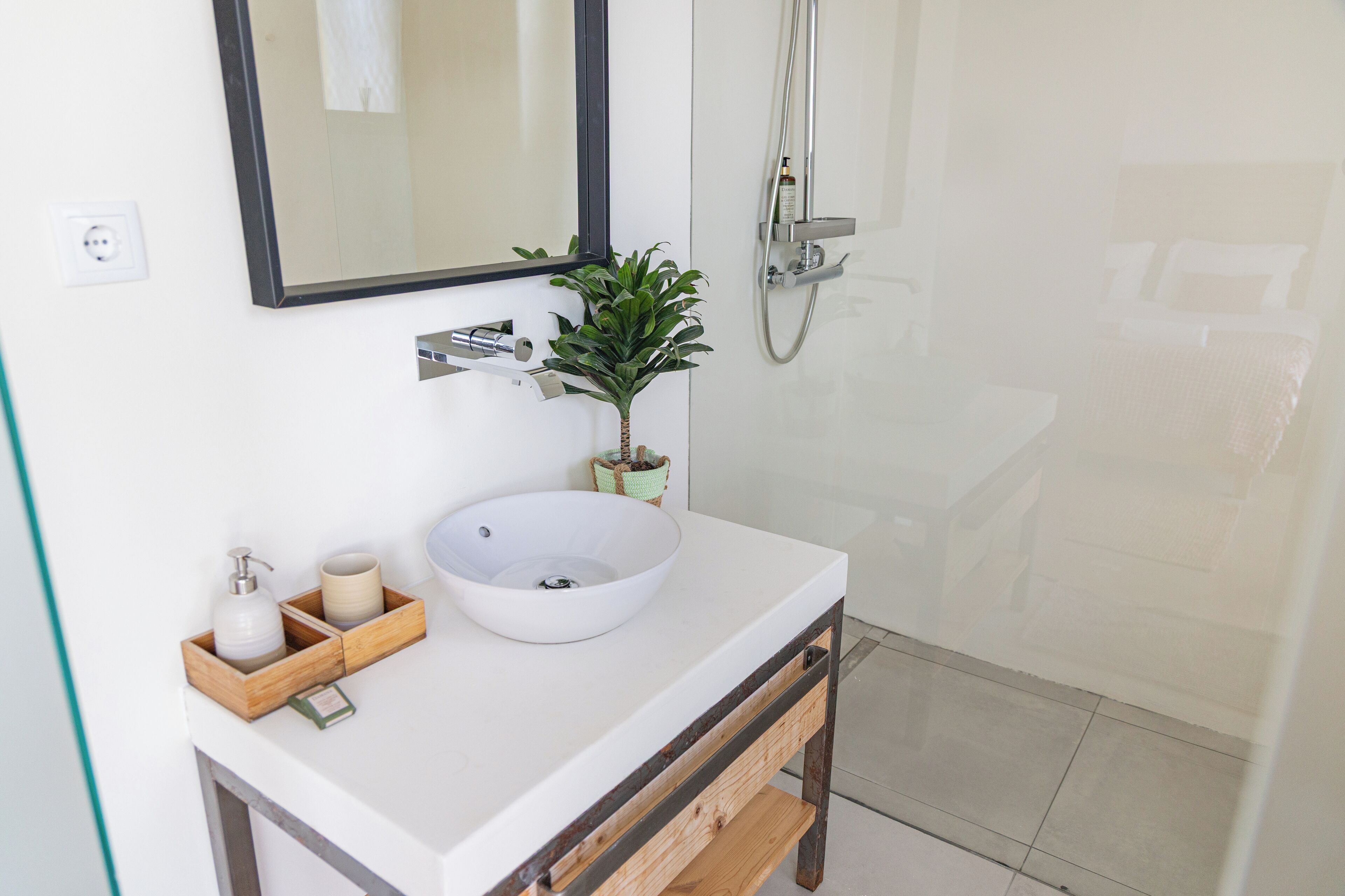 Un baño con lavamanos blanco, espejo, ducha y planta en maceta.