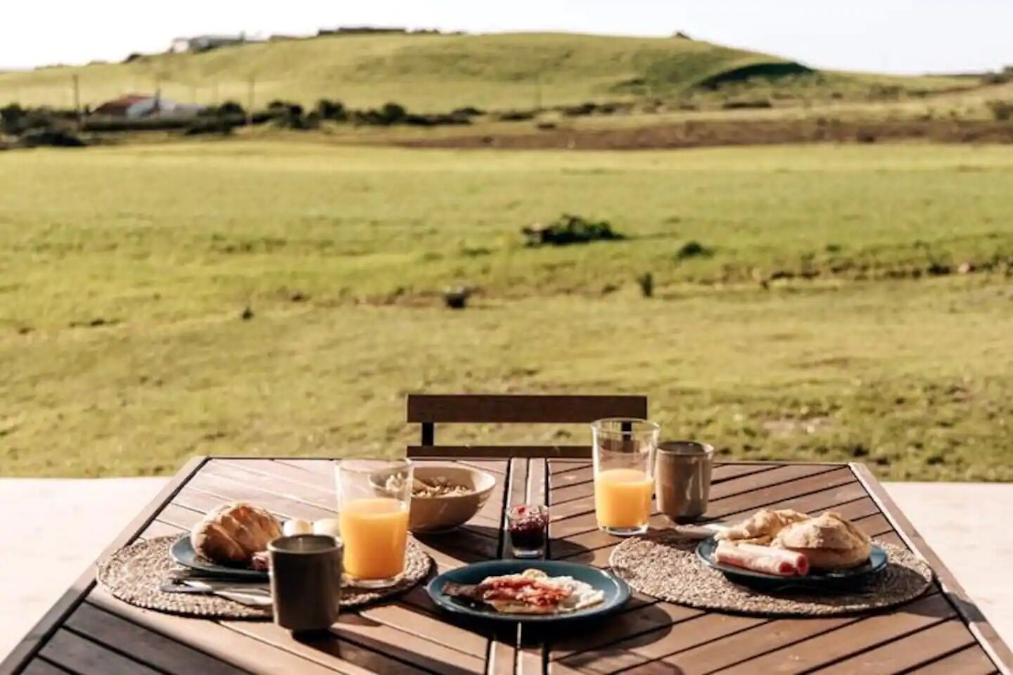 A table set outdoors with breakfast items and drinks, overlooking a grassy field.