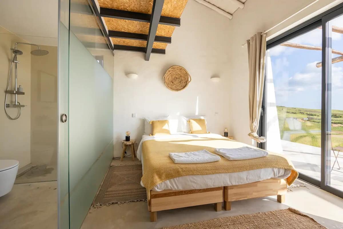 A bedroom with a bed, bedside table, lamp, and a view of the countryside.