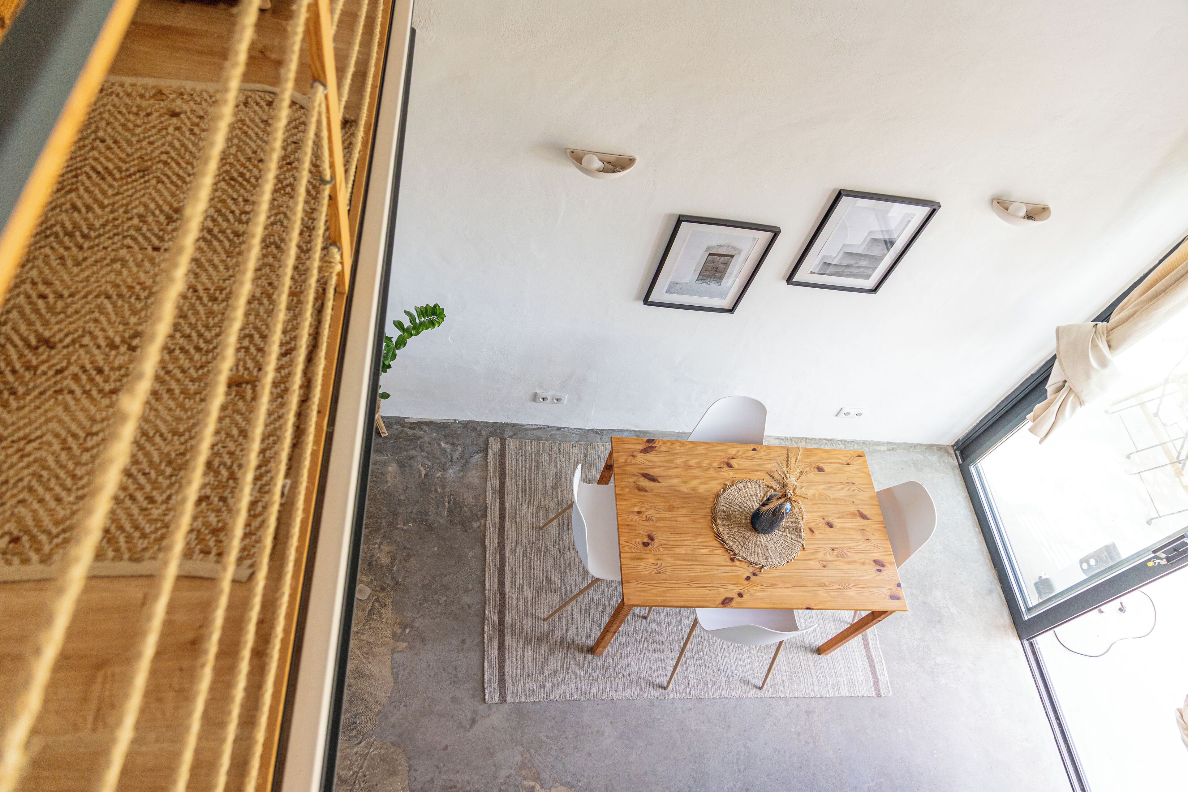 Een eetgedeelte met een houten tafel, witte stoelen en een geweven placemat. Er hangen twee ingelijste foto's aan de muur en er staat een plant op de vloer.