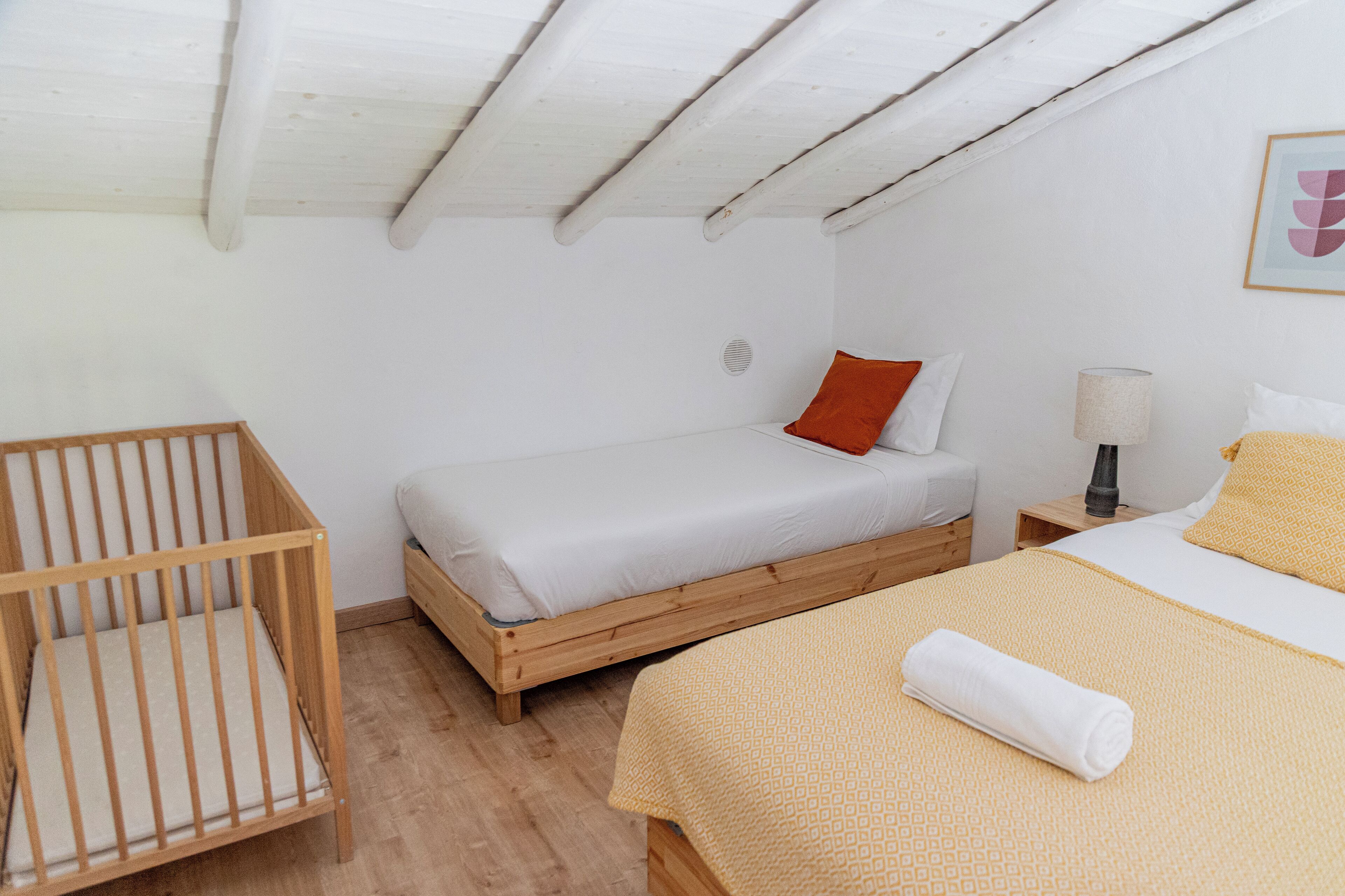 Een slaapkamer met een houten babybedje, een enkel bed met een gele sprei, een nachtkastje met een lamp en een witte muur met een lijstwerk.