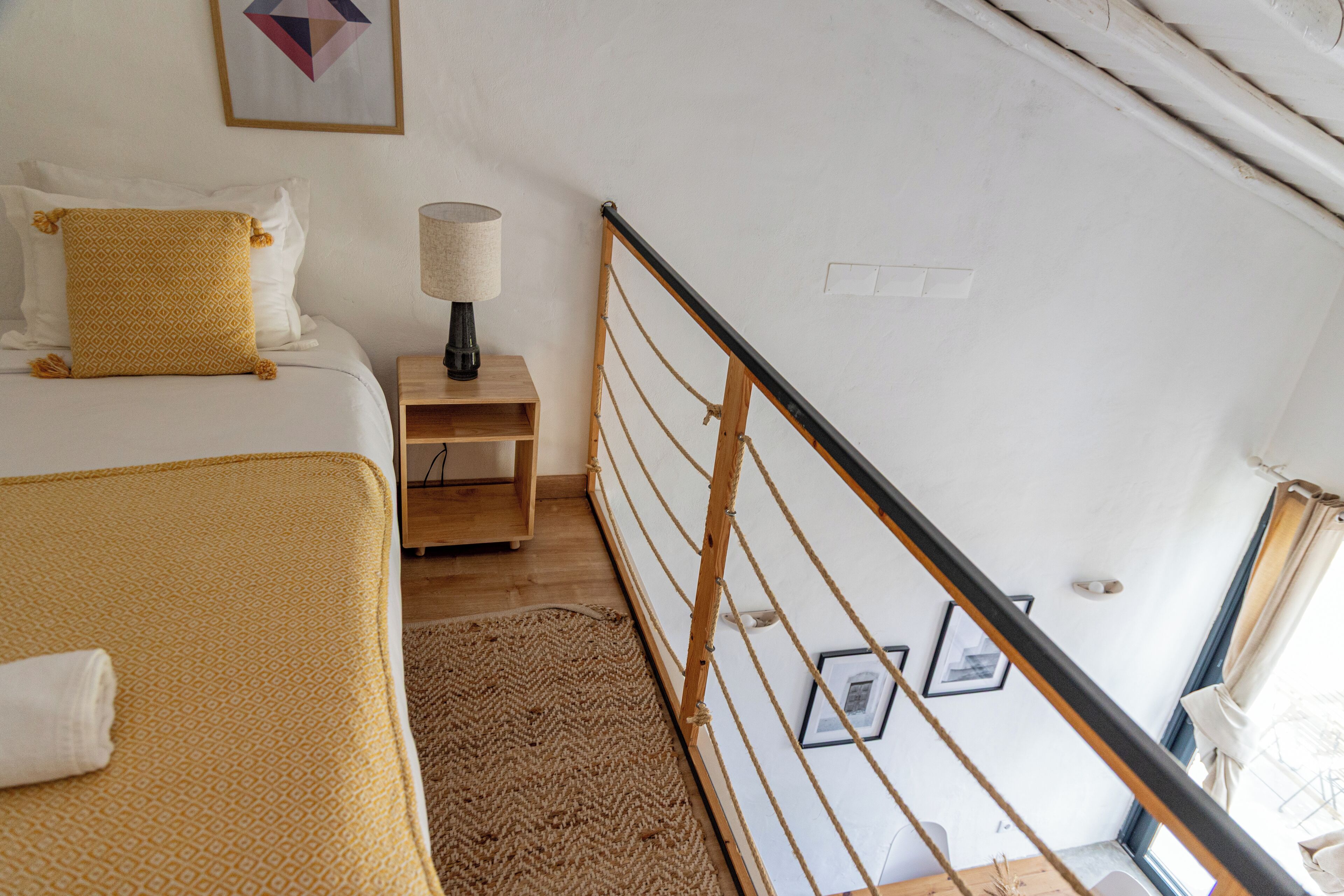 A bedroom with a bed, a nightstand, a lamp, a wooden floor, a railing, and a window with curtains.