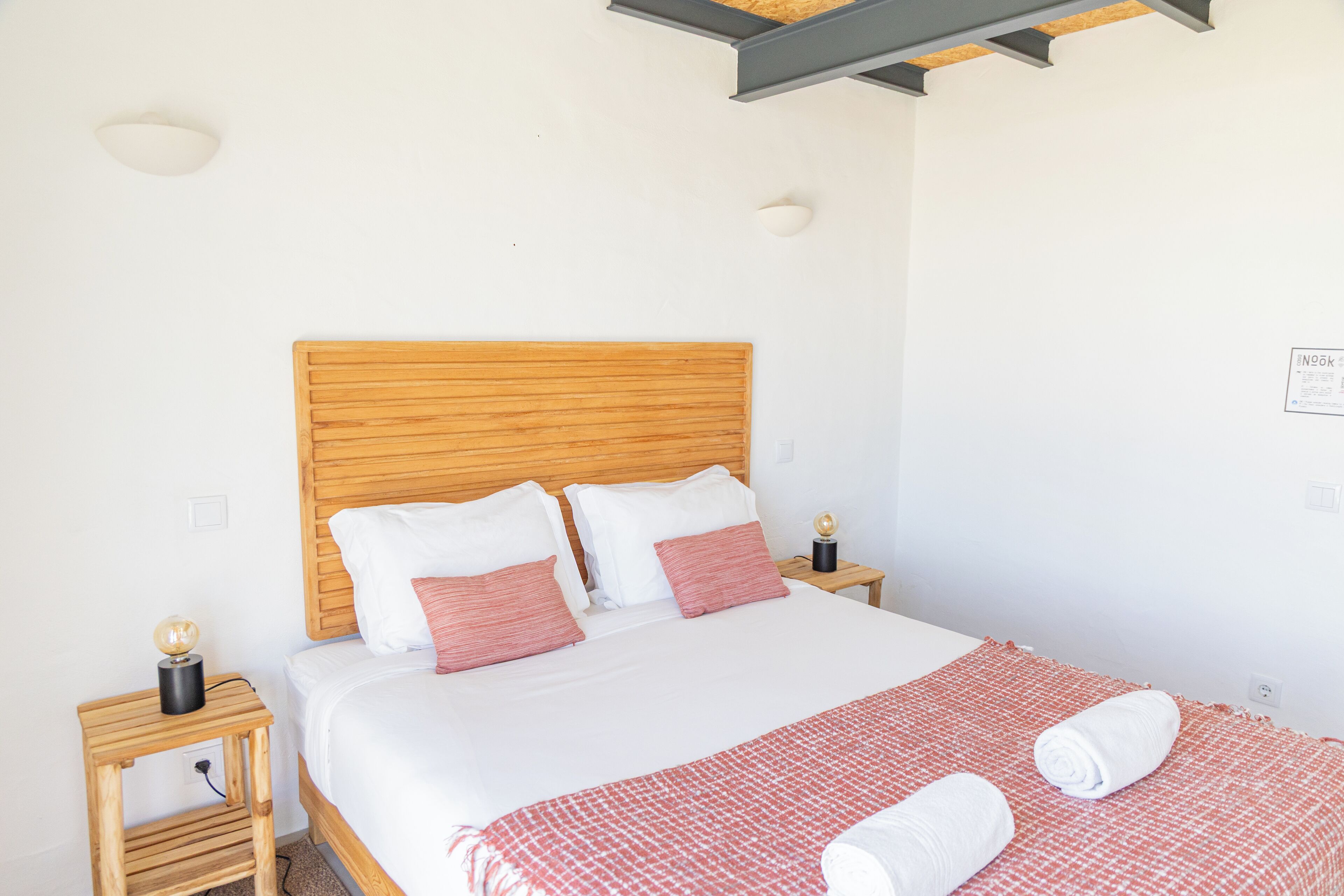 Uma cama bem arrumada com cabeceira de madeira, roupa de cama branca e um cobertor vermelho estampado. Há duas mesas de cabeceira, com luminárias e um globo em uma delas.