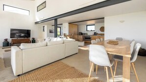 Een moderne woonkamer met een grote bank, een eettafel met stoelen, een televisie en een keuken in de achtergrond.