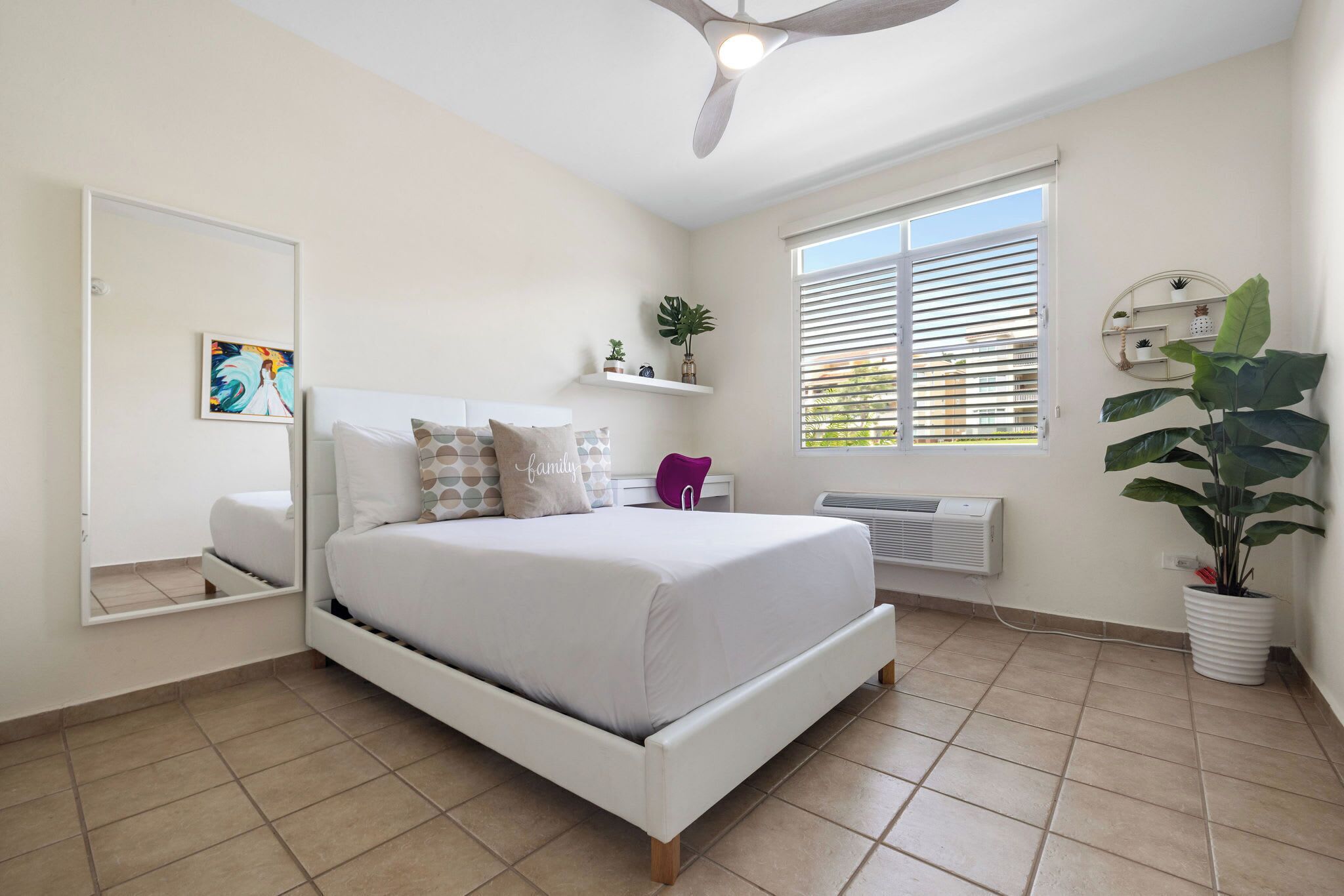 Une chambre à coucher avec un lit, un ventilateur de plafond, une fenêtre avec des stores, un miroir et une plante en pot.