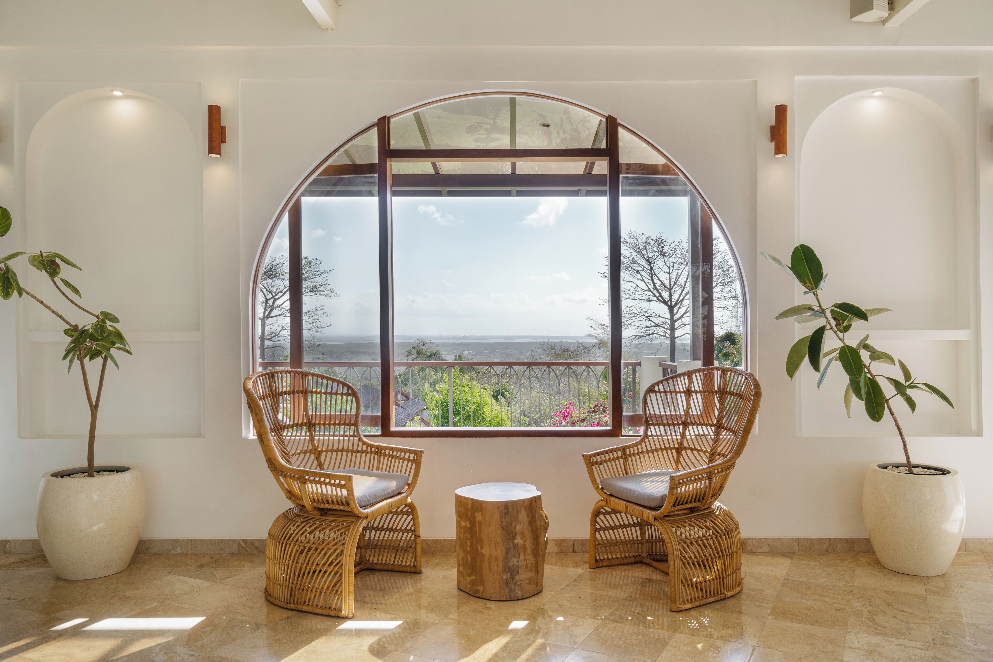A room with a large arched window, two wicker chairs, a small wooden table, and potted plants.