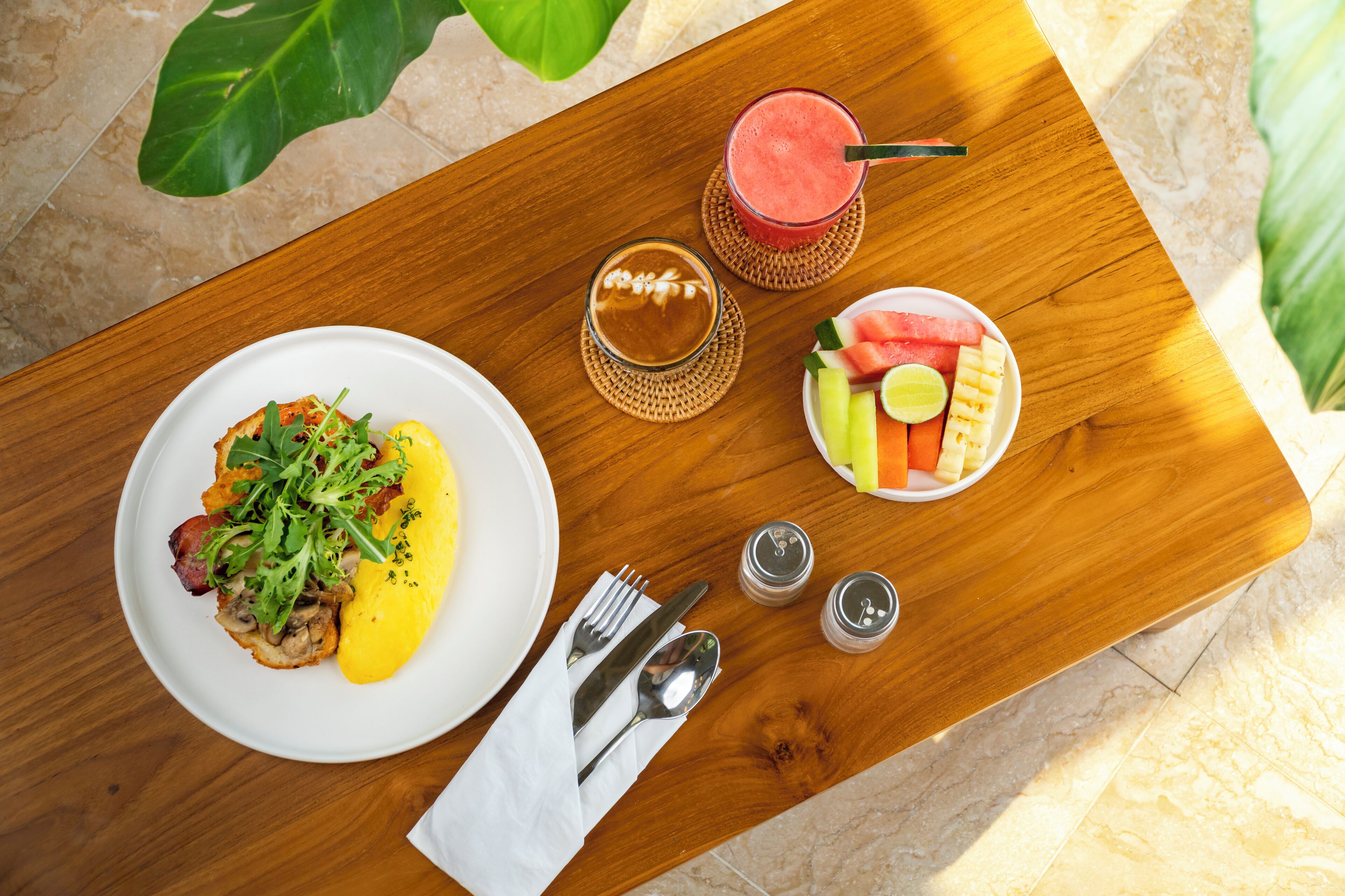 A wooden dining table with a plate of food, a glass of iced coffee, a glass of smoothie, and a plate of fruit.