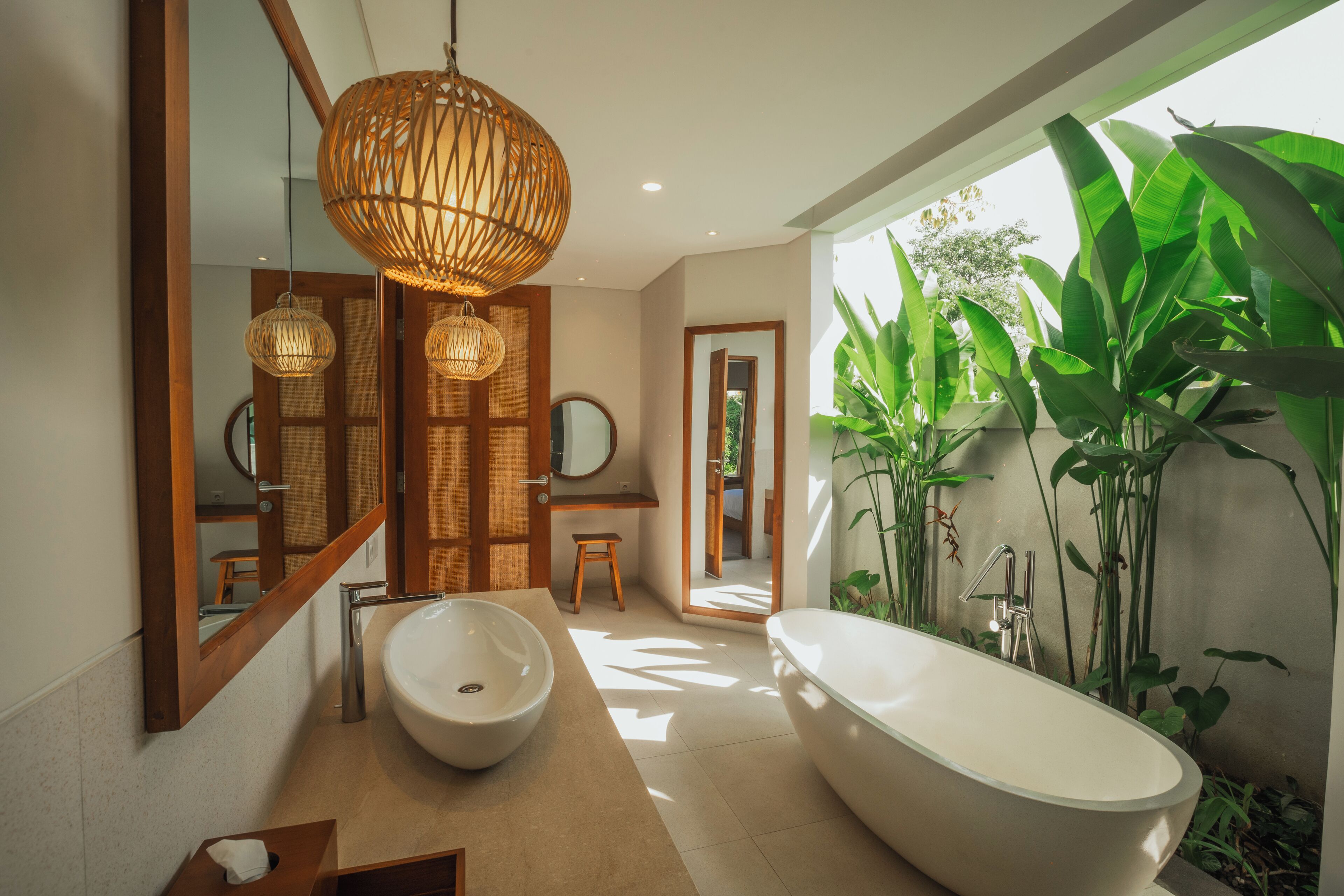 Une salle de bain moderne avec une baignoire sur pieds, un grand miroir et un lavabo avec un receveur apparent. On y trouve des luminaires en osier et une vue sur la verdure à l’extérieur.