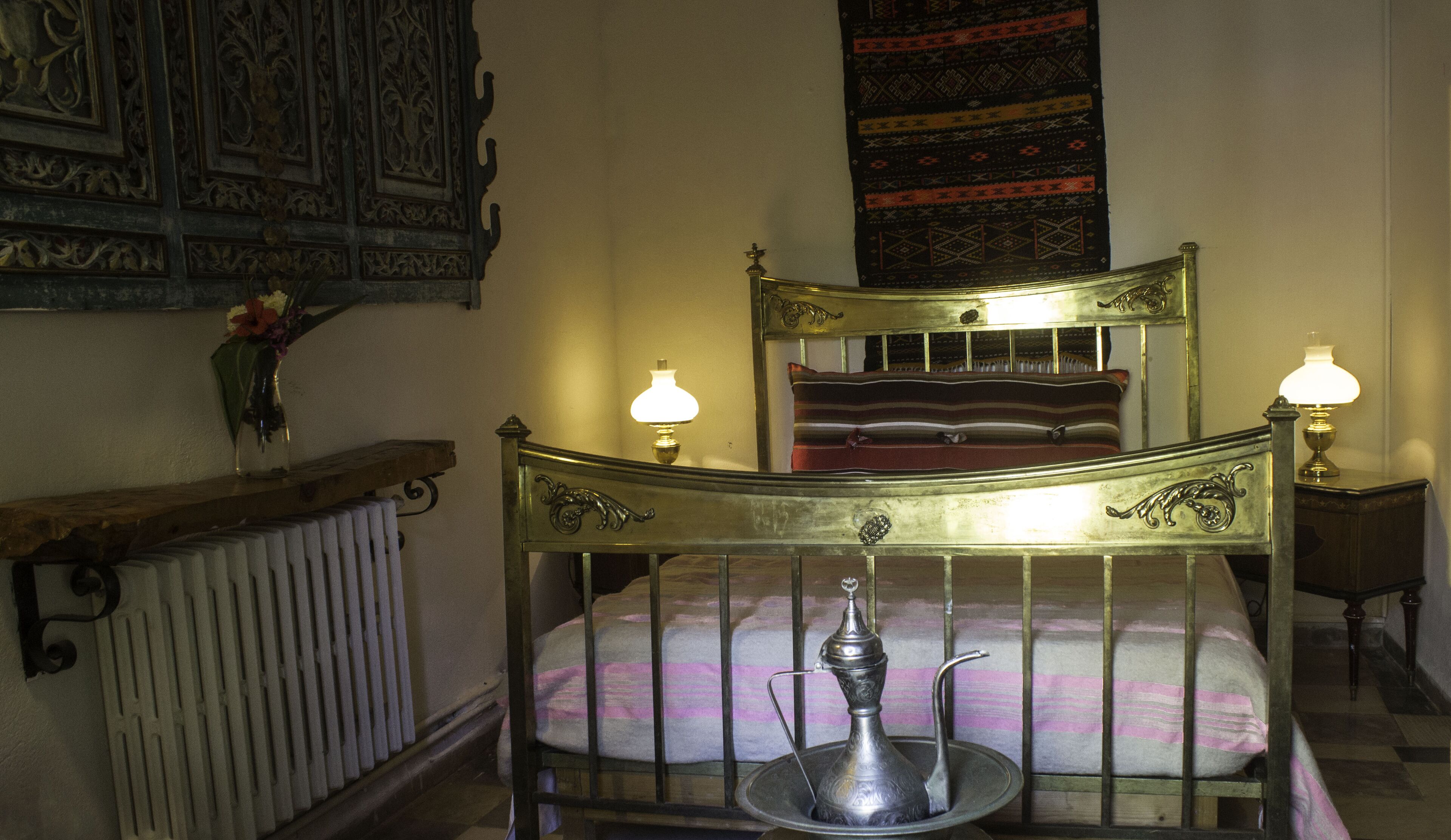 Una cama tradicional con ornamentales cabecera y piecera de metal, dos lámparas de noche, un radiador y un tapiz decorativo en la pared.