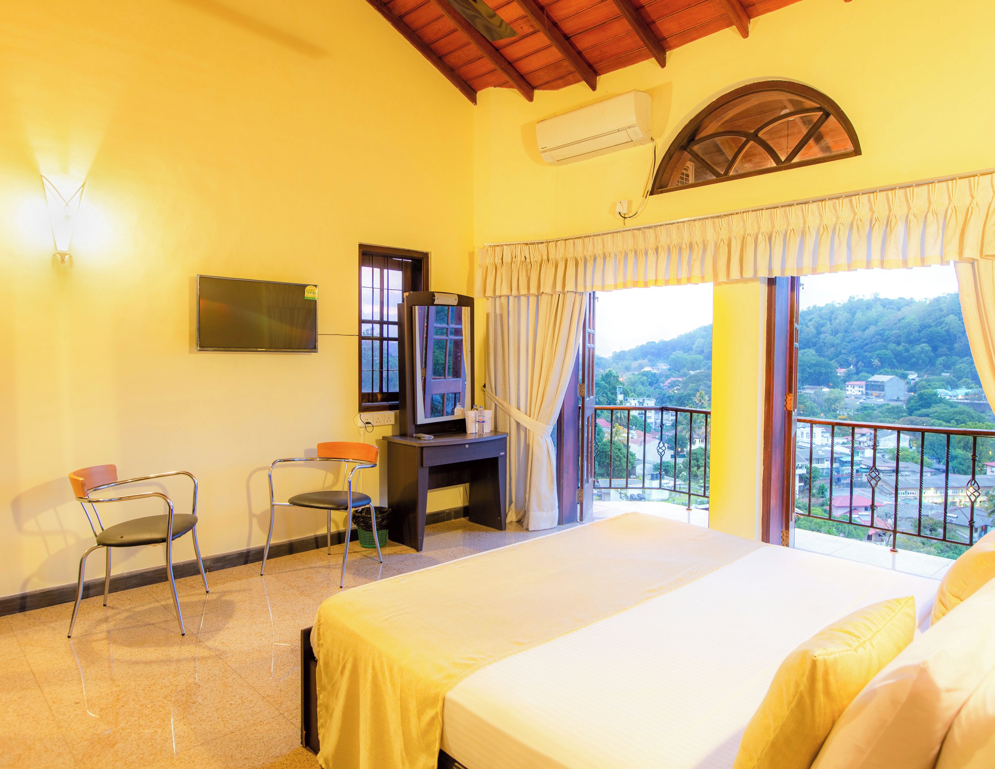 A hotel room with a bed, a desk with a computer, a chair, a TV, and a balcony with a view of the city.