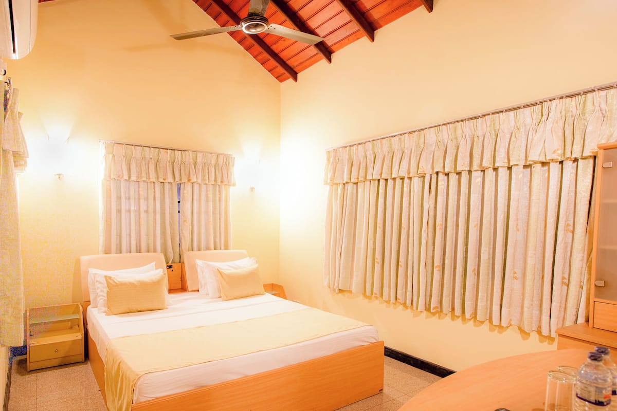 Ein Hotelzimmer mit Bett, Nachttischen, Deckenventilator, Fenster mit Vorhängen sowie einem kleinen Tisch mit Fernbedienung und Wasserflasche.