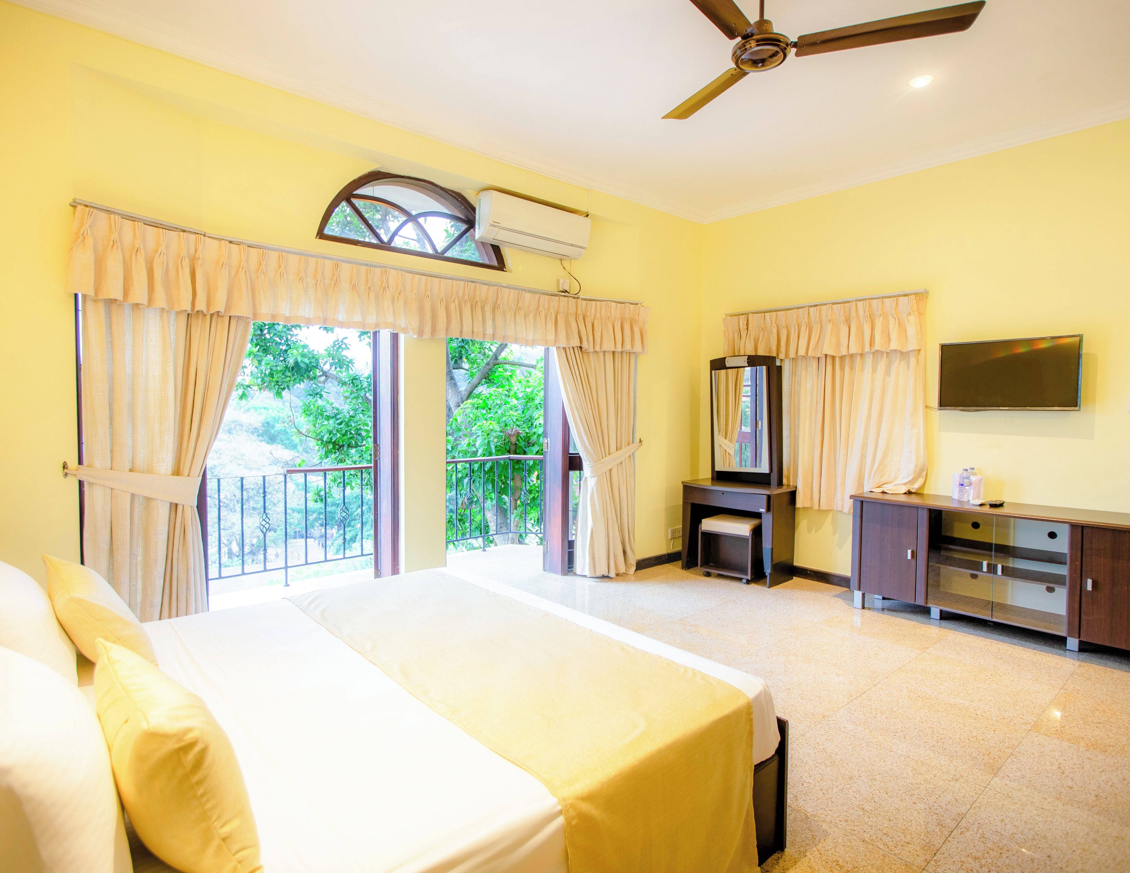 A hotel room with a bed, a vanity with a mirror, a television, and a ceiling fan.