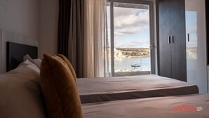 A hotel room with a bed, a large window with a view of a body of water and a distant landscape, and a wardrobe. - Seaside Magic From a Stunning Apartment (St. Paul's Bay)