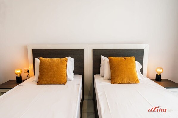 Deux lits jumeaux avec du linge de lit blanc et des oreillers couleur moutarde, chacun étant muni d’une lampe de chevet.