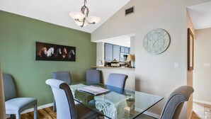 A dining area with a glass table and grey upholstered chairs, a chandelier, a clock, and a kitchen in the background.