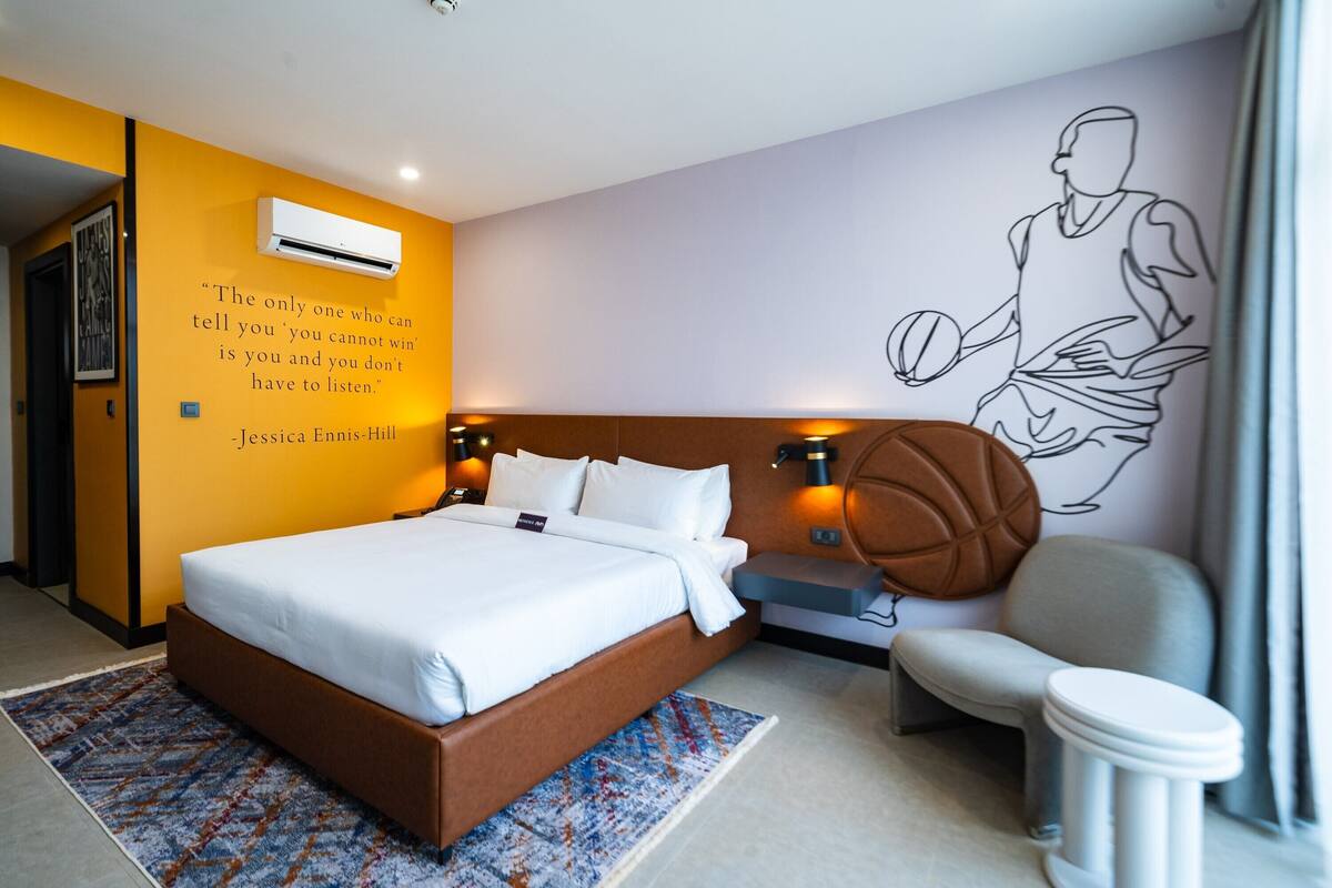 A modern hotel room with a large bed, a basketball-themed wall mural, a small seating area, and a quote by Jessica Ennis-Hill.