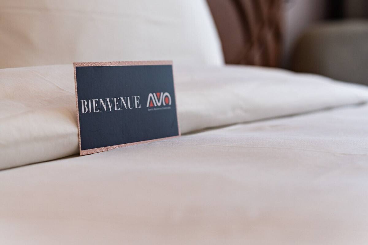 A hotel room with a bed, a welcome sign, a wall-mounted lamp, and a decorative wall piece.