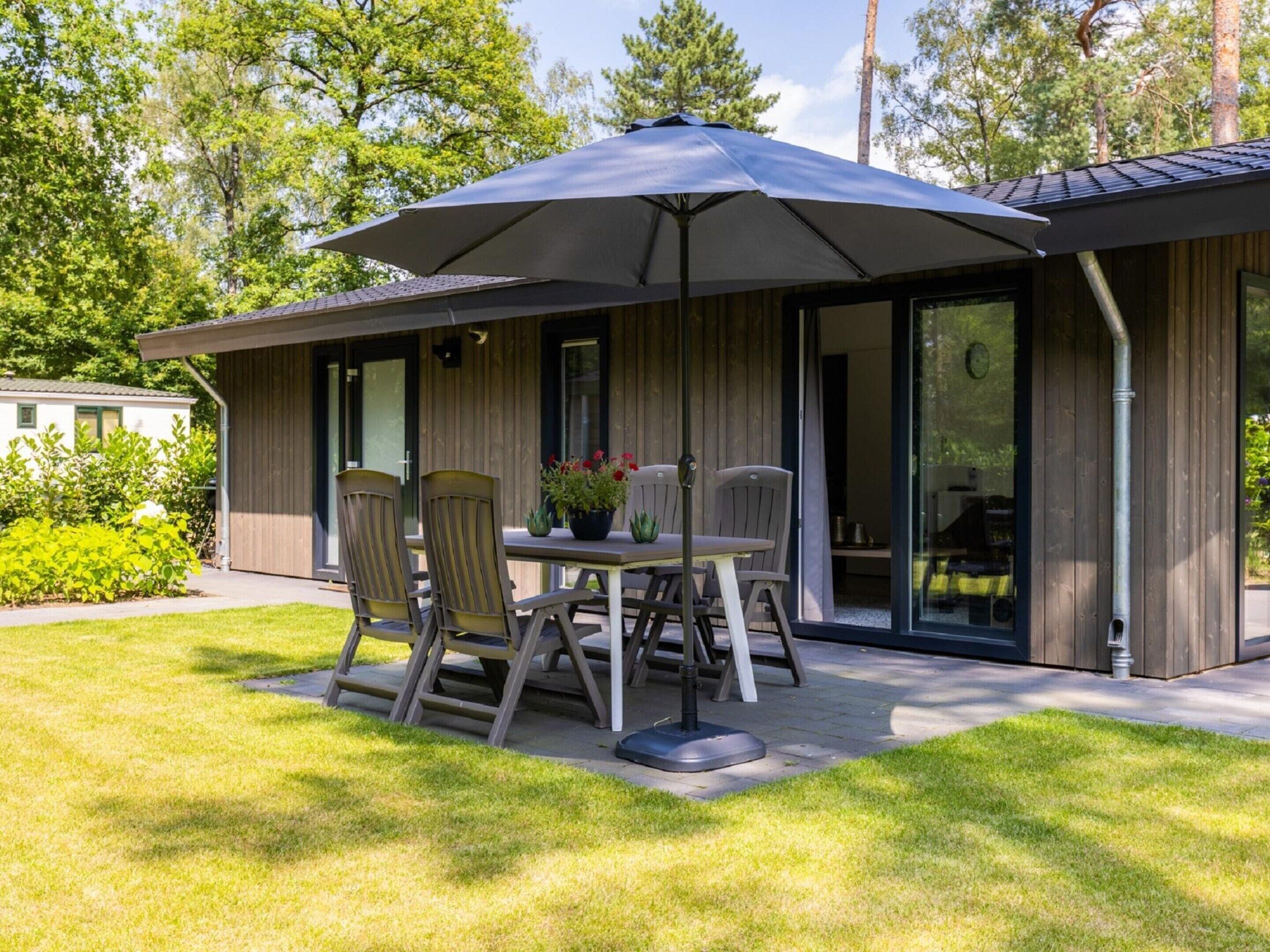 Un patio moderno al aire libre con una mesa, sillas, un paraguas grande y un edificio de madera con puertas de vidrio.