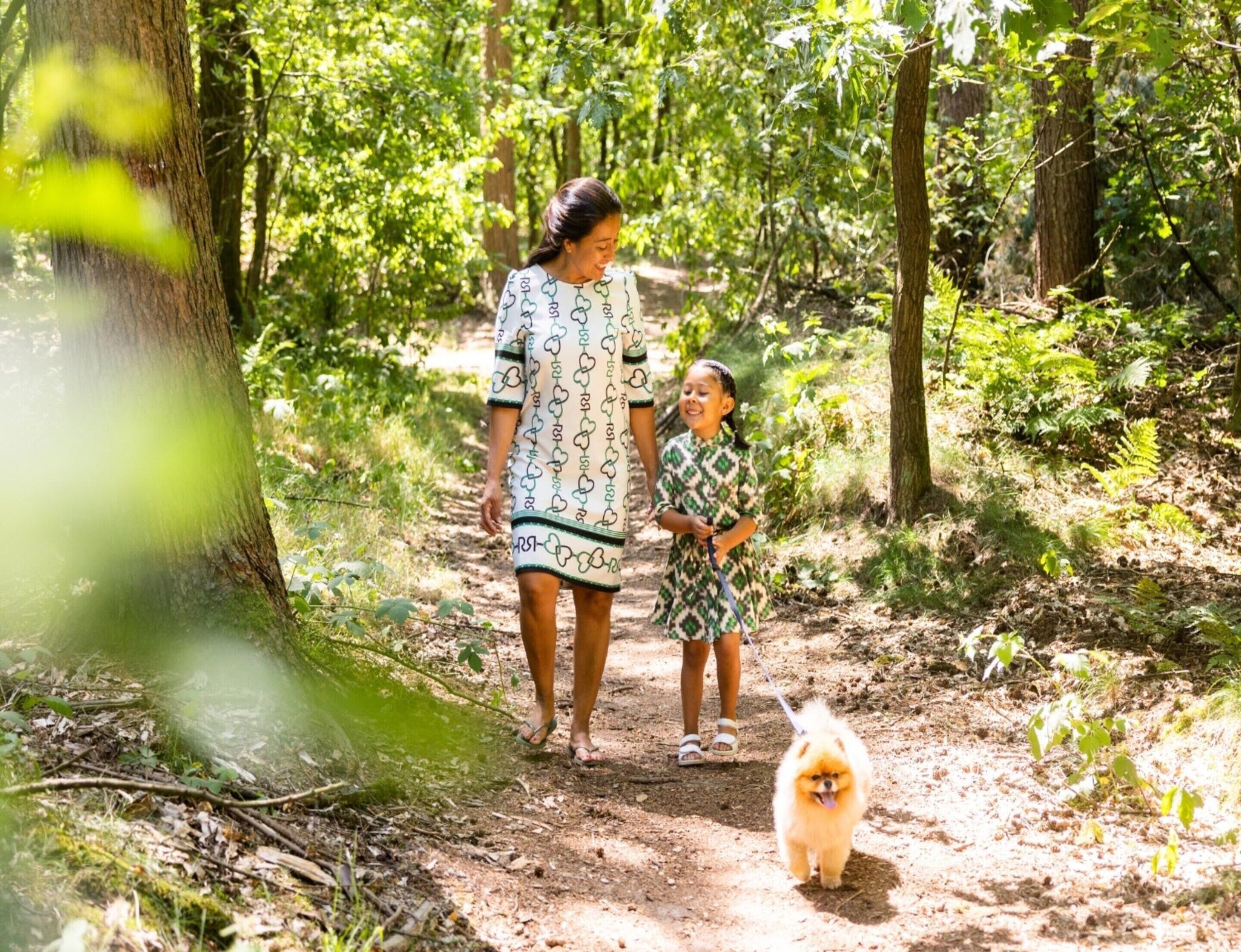 A woman and a child walking a small dog on a dirt path in a wooded area.