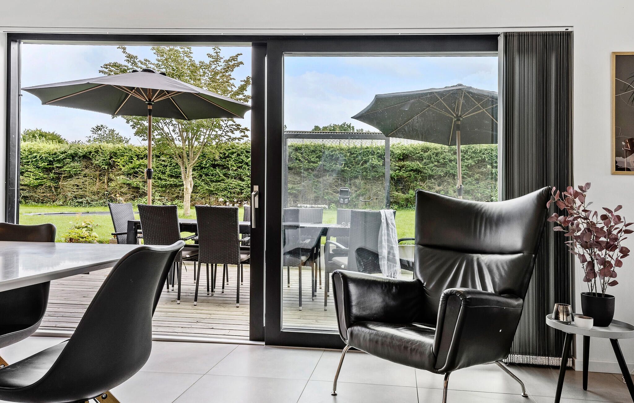 Un espacio moderno para comer al aire libre con puerta de cristal que da a un jardín, con una mesa, sillas y un gran paraguas.