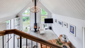 Spazioso loft con soffitto alto, una stufa a legna in legno nero, un lampadario moderno, una TV a schermo piatto e un'area pranzo con vista sull'esterno.