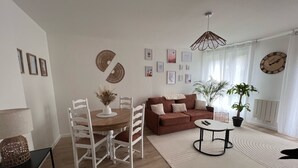 A modern living room with a brown sofa, a round coffee table, and a dining table with chairs. There are several framed pictures on the walls and a large window with sheer curtains.