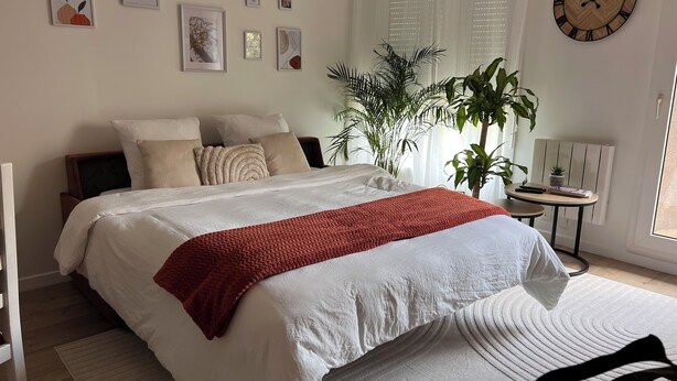A neatly made bed with a white and red blanket, a bedside table with a plant, and a clock on the wall.