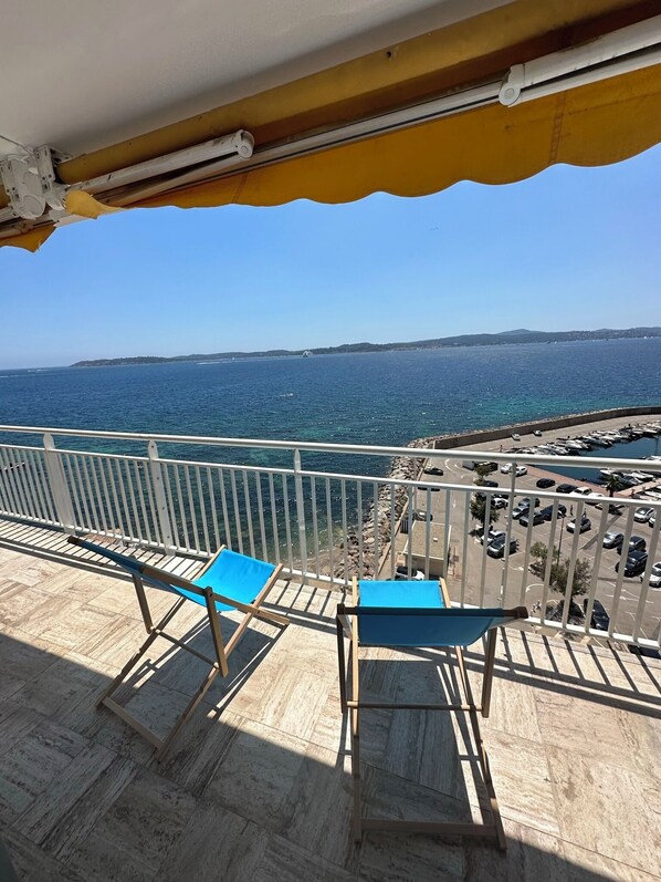 A balcony with two blue chairs and a view of the sea and a parking area.