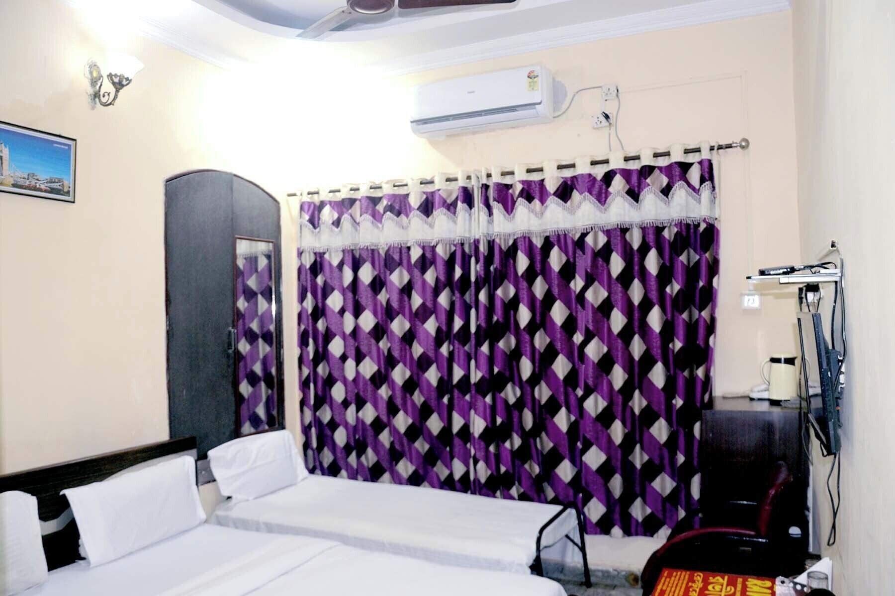 A hotel room with a bed, a desk, an air conditioning unit, and a curtain with a diamond pattern.