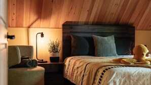 A cozy bedroom with a wooden ceiling, a bed with a patterned bedspread, a bedside lamp, a chair, and a potted plant.