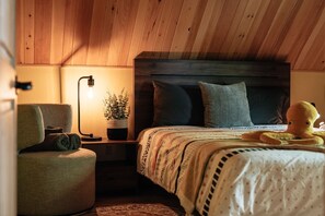 Ein gemütliches Schlafzimmer mit Holzboden, einem Bett mit gemustertem Überwurf, einer Nachttischlampe, einem Sessel und einem Topf mit Pflanze.