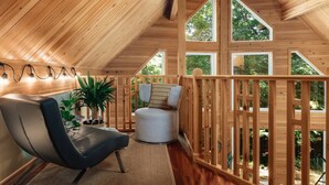 A cozy cabin interior with a wooden ceiling, a cushioned chair, a small table with a plant, and a view of the outdoors through large windows. - SPA & Nature | Chalet AYUR (La Minerve)
