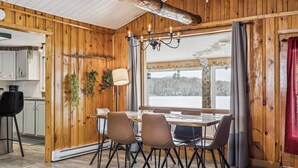 A wooden interior with a dining table and chairs, a chandelier, and a view of a snowy landscape through a large window. - Waterfront - Spa | Havre de l'Auror | Harry Potter (Outaouais)