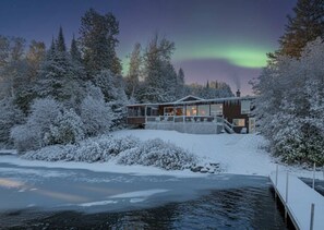 Een besneeuwde lodge met een steiger die uitsteekt in een bevroren meer, omringd door dichte bomen.