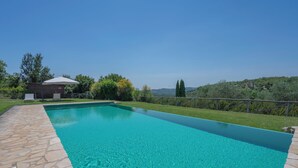 Une piscine rectangulaire à l’eau d’un bleu limpide, entourée d’une terrasse en pierre et d’une pelouse agrémentée d’arbres et, au loin, de collines.