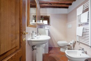 Bathroom features a pedestal sink, toilet, bidet, and mirror with a wooden frame, set against a backdrop of small tiles.