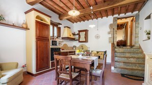 Un coin repas avec une table et des chaises en bois, une cuisine équipée d’une cuisinière et de placards, et un escalier en pierre menant à un autre niveau.