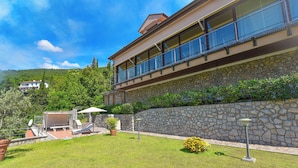 A two-story building with a stone facade, a balcony with glass railings, and a well-maintained lawn with outdoor furniture. - Apartment Vosilla 2 (Opatija)