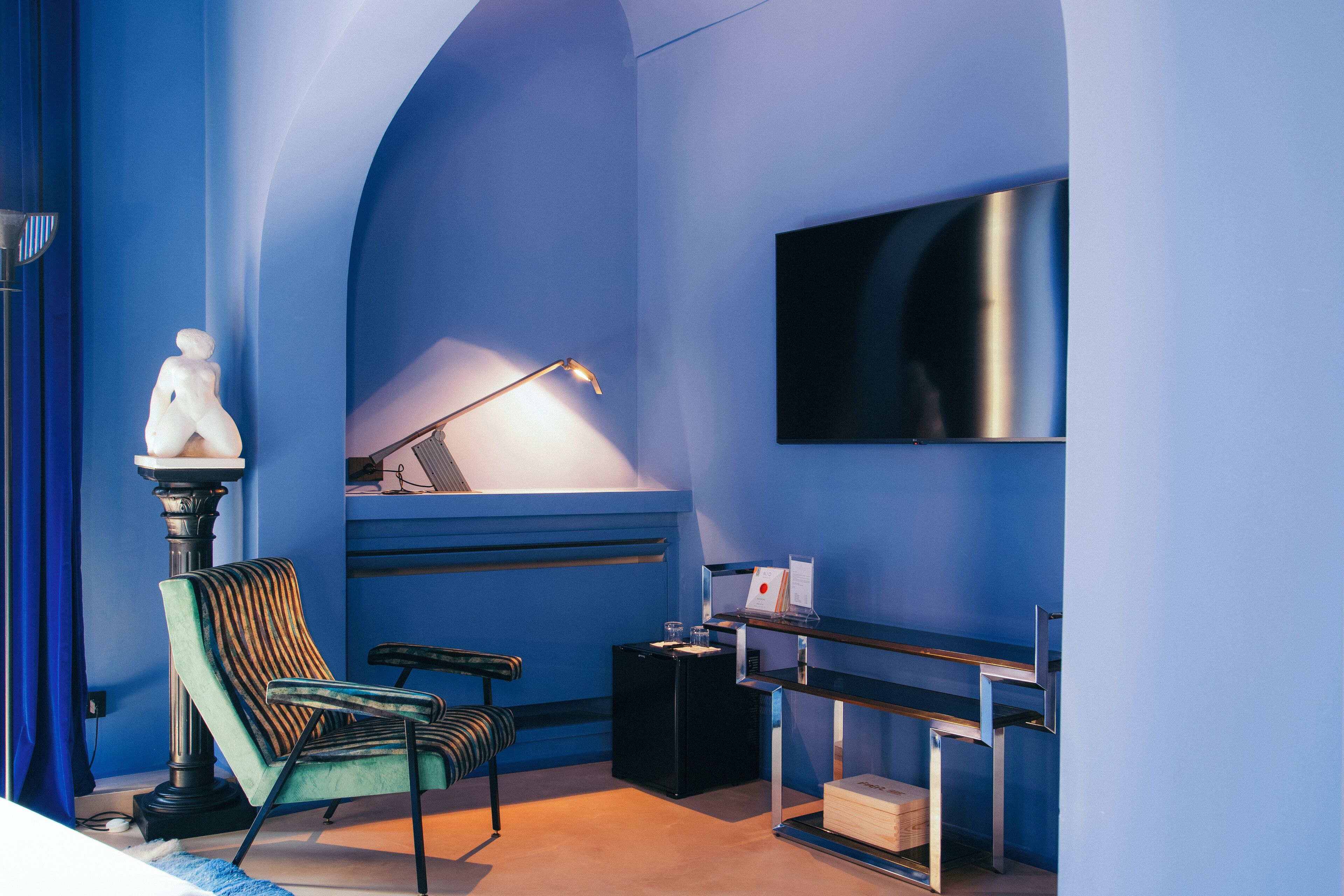 A room with a blue wall, a mounted TV, a desk with a lamp, and a chair.