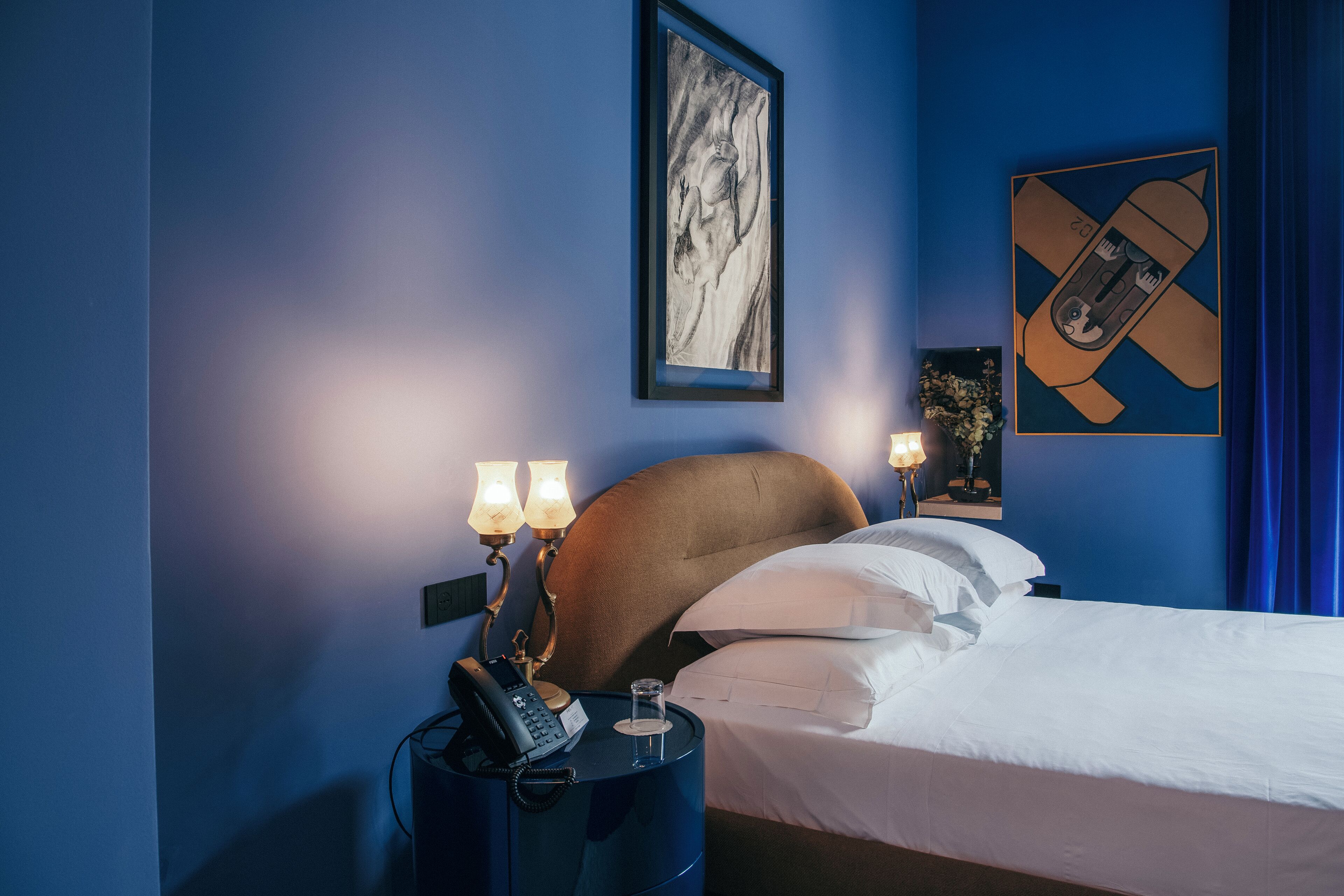 Une chambre d’hôtel avec un grand lit, une table de chevet avec un téléphone et des lampes, et des œuvres d’art accrochées aux murs.