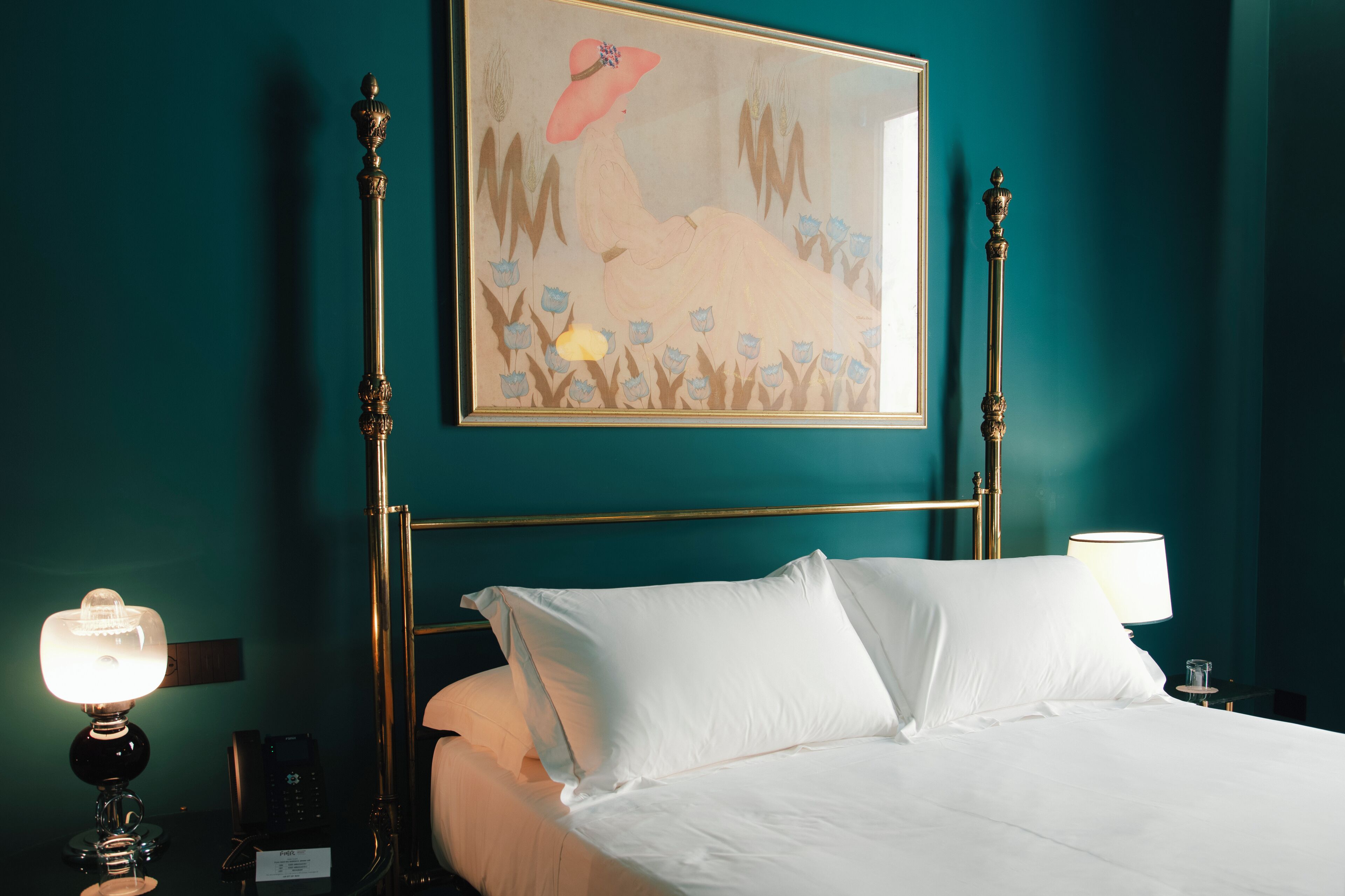 Quarto de hotel com paredes verde-água, cama com roupa de cama branca, quadro emoldurado sobre a cama, abajur e telefone na mesa de cabeceira.