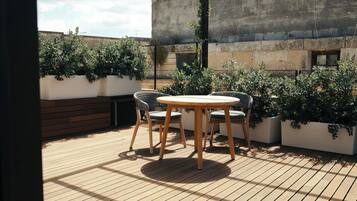 Um deck de madeira com uma mesa redonda e cadeiras, cercado por plantas em vasos e uma parede de concreto.