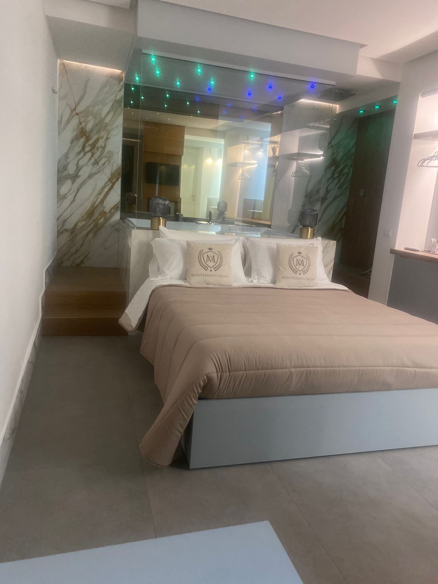 Une chambre à coucher moderne avec un mur en marbre, un lit recouvert d’une courtepointe beige, une coiffeuse avec un miroir et un escalier menant à un niveau supérieur.