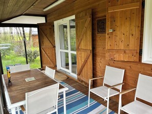 A wooden cabin with a dining area featuring a table and chairs, large windows, and a view of the surrounding greenery. - Chalet et Jardin Dans un Domaine Privé (Arzal)