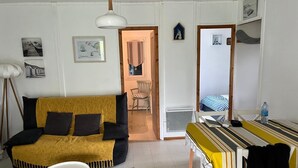 Ein kompakter Wohnbereich mit einer Couch, einem kleinen Tisch und einem Esstisch. Ein Durchgang führt in einen weiteren Raum mit einem Bett.