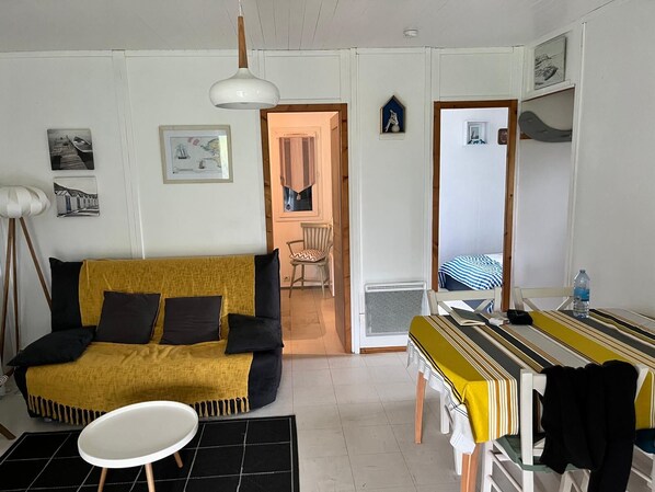 Un espacio habitable compacto con sofá, una mesita y una mesa de comedor. Se ve un acceso a otra habitación con una cama.